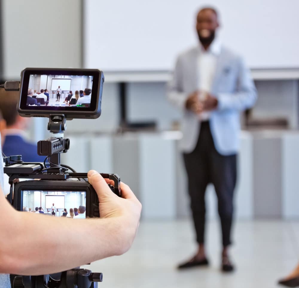 Vidéaste filmant un homme qui donne une présentation devant un groupe, affiché sur l'écran de la caméra